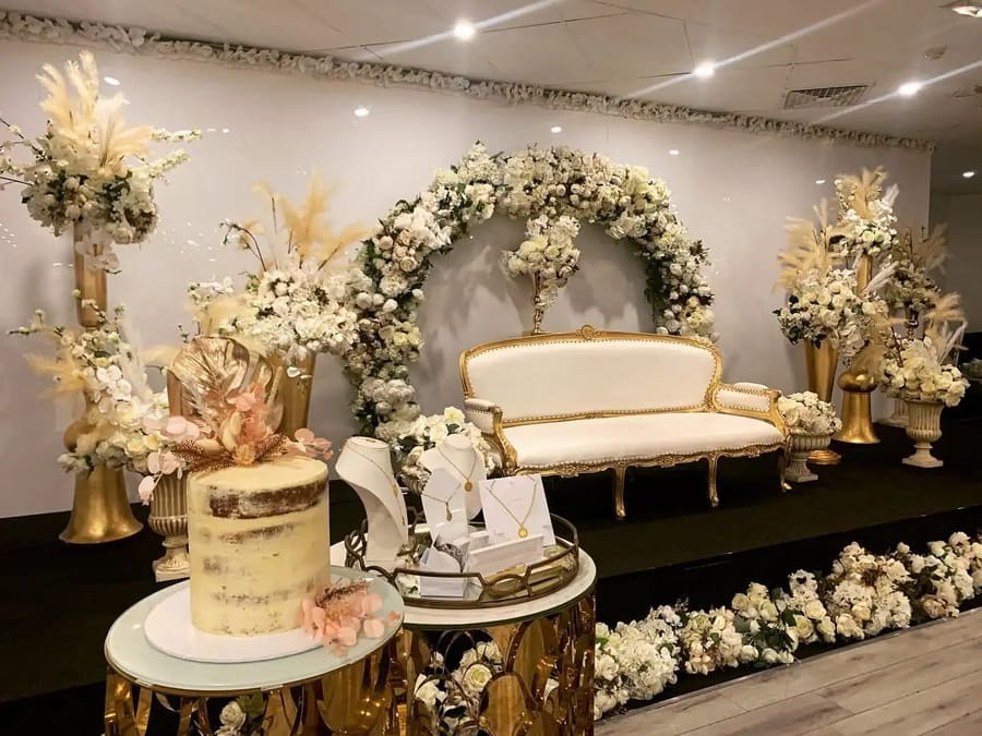 Gold-trimmed lounge under a white floral arch, with a semi-naked cake in front